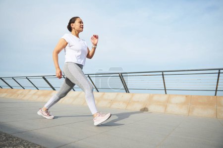 Foto de Mujer madura haciendo footing matutino en el puente de la ciudad. Activa joven mujer atlética adulta en camiseta blanca y polainas grises, corriendo durante la maratón de concientización, desafiante. Gente activa. Deporte - Imagen libre de derechos