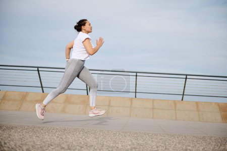 Foto de Retrato lateral de una mujer corriendo por el puente de la ciudad, haciendo footing matutino, haciendo ejercicio al aire libre. Atlética hembra quemando calorías y perdiendo peso mientras corre trotando corriendo en el paseo marítimo - Imagen libre de derechos