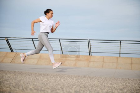 Frau läuft auf einer Stadtbrücke ins Freie. Aktive Sportlerinnen genießen ein morgendliches Training, Joggen. Gesunder Lebensstil. Sportliche Person in Sportbekleidung, die im Freien trainiert, weißes T-Shirt