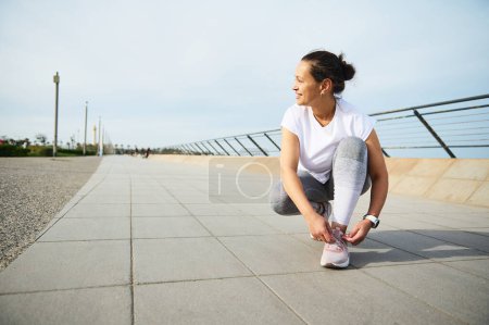 Young woman jogger tying laces on her pink sneakers, smiling looking away, getting ready for morning jog in the city outdoors. Running and jogging. Awareness running marathon concept. Sport. Endurance