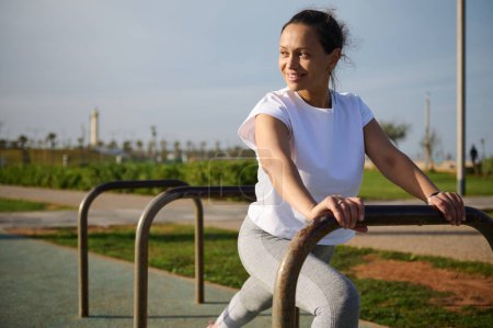 Foto de Mujer atlética joven determinada en camiseta blanca y polainas grises, sonriendo mirando a un lado, calentando su cuerpo, haciendo embestidas para estirar los músculos de las piernas, haciendo ejercicio en el campo deportivo urbano al aire libre - Imagen libre de derechos