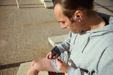 Selective focus on a sportsman athlete man in gray activewear, holding earphones and smart mobile phone, relaxing after outdoors workout, checking fitness app, listen music. People. Sport. Technology
