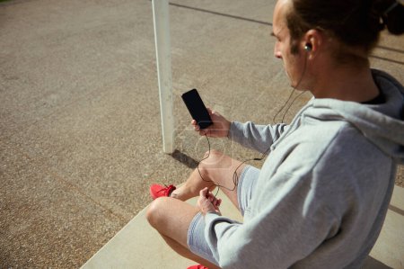 Close up of a sportsman wearing earphones, using smartphone with blank black mockup digital touch screen, browsing new fitness app on mobile phone while resting and checking results of outdoor workout