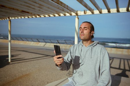 Young Caucasian man in gray sportswear, wearing earphones, holding smart mobile phone, listening to music while resting after workout, sitting on a stone bench against Atlantic beach background