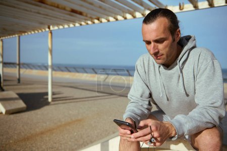 Caucasian active young man holding mobile phone, browsing web sites or fitness app, scrolling newsfeed while checking social media content, sitting on bench in the Atlantic promenade on warm sunny day