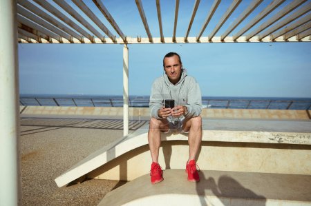 Junger Mann in Sportkleidung, überprüft Fitness-App, während er sich nach dem Outdoor-Training ausruht, sitzt auf einer Bank mit Zelt. Menschen. Sport. Technologie. Lebensstil. Online-Kommunikation
