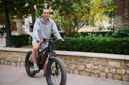 Full length portrait of a handsome Caucasian man commuter renting electric bike, smiling looking at camera while riding in the city. Bike sharing city service
