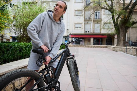 Jeune homme ajuste la hauteur du nouveau vélo à batterie, vélo électrique avant de le sortir pour la première fois. Beau type louant une moto électrique à batterie, en ville. Transports écologiques