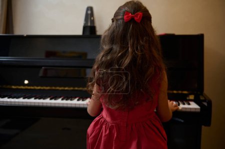 Vista desde la parte posterior de una niña con elegante vestido rojo, sentada en el forte del piano, interpretando melodía clásica, sintiendo el ritmo de la música, tocando las teclas de marfil blanco y negro y ébano del piano de cola