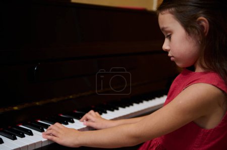 Auténtico retrato de niña linda caucásica en elegante vestido rojo, sentada en el forte del piano, interpretando melodía clásica, sintiendo el ritmo de la música, tocando las teclas de marfil y ébano del piano de cola