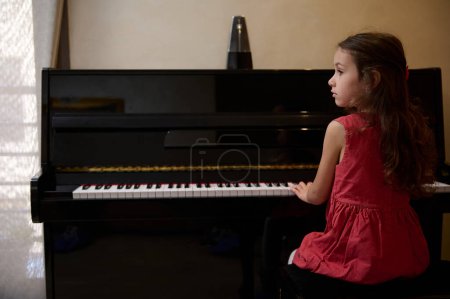 Confident portrait of an inspired beautiful talented little child girl learning music, performing the rhythm of classical music while playing piano, putting fingers on keys and dreamily looking away