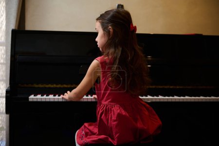 Authentic Caucasian little child girl musician pianist with beautiful long hair, dressed in elegant red dress, putting fingers on piano keys, enjoying the performance of classical music. Copy ad space