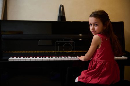 Auténtica niña con un elegante vestido rojo, mirando a la cámara, sentada al piano e interpretando una melodía clásica en piano de cola. Adorable niña tocando el piano en casa. Copiar espacio anuncio