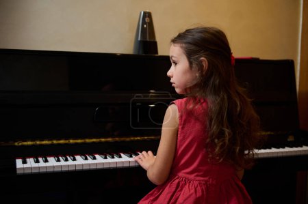 Chica de escuela caucásica confiada en vestido rojo, sentada en una silla y realizando composición musical clásica, tocando las teclas y sintiendo ritmo mientras toca el piano de cola en casa. Música. Gente.