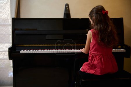Back view of a little child girl pianist in stylish red dress, playing grand piano at home. Talented kid, musician composing a melody, putting fingers on black and white piano keys. Copy space