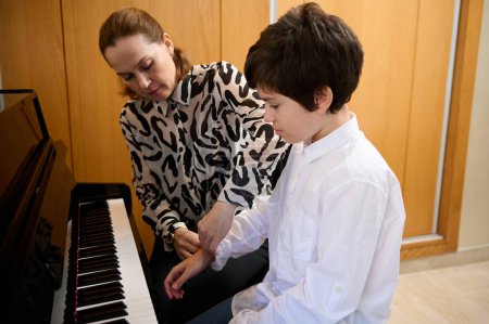 Handsome teenage boy in white shirt, taking piano lesson, passionately playing the keys under her teacher's guidance, feeling the rhythm of music. Musical education and talent development in progress