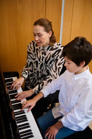 Retrato vertical de un adolescente auténtico que tiene clase de música en casa, sentado cerca de su maestro pianista, tocando el piano de cola, creando composición musical y sintiendo el ritmo de los sonidos