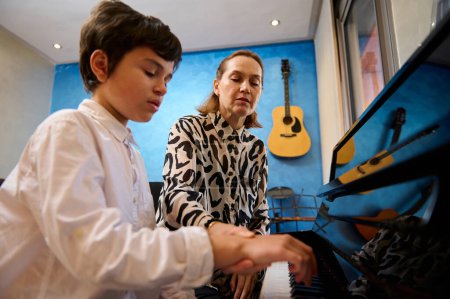 Middle aged female pianist, music teacher teaching piano and music lesson to a teenage boy in white casual shirt, sitting together at wooden piano. People. Lifestyle. Education. Hobbies and leisure