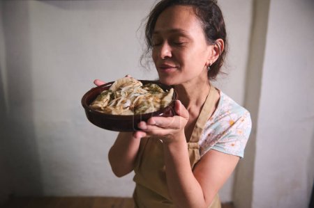 Jeune femme au foyer dans le tablier du chef beige, tenant un plat d'argile avec des boulettes maison fraîchement cuites Varenyky avec de la purée de pommes de terre et des oignons rôtis. Cuisine et culture ukrainienne traditionnelle