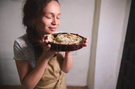 Authentique portrait d'une belle jeune femme brune en tablier de cuisine beige, souriant, savourant l'arôme de boulettes fraîchement cuites avec purée de pommes de terre et oignons rôtis, servi dans un plat d'argile