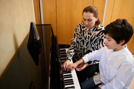 Authentic portrait of a female pianist musician holding the hands of a teenage boy, showing the true position of finger on piano keys, explaining the piano lesson during individual music class at home