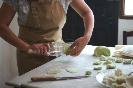 Close-up of a housewife using glass wine bottle as rolling pin, rolling out green spinach dough for making dumplings, standing at kitchen table sprinkled with flour in the rural house kitchen