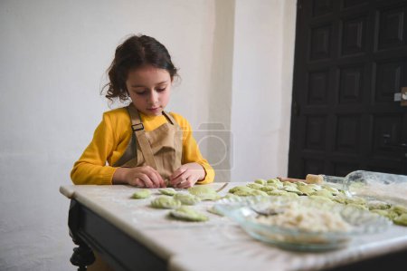 Caucasian charming child girl making dumplings in the home kitchen, dressed in yellow sweater and beige chef's apron. People. Culinary. Child learning cooking. Childhood. Domestic life