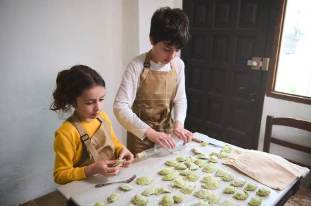 Cute children in the rural kitchen, rolling out dough with a wine bottle, sculpting dumplings with mashed potatoes filling. Cooking homemade vegetarian dumplings, Italian ravioli or Ukrainian varenyky