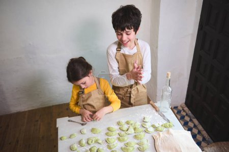 View from above of two diverse kids, a boy and a girl preparing family dinner, standing at floured kitchen table and modeling dumplings or Ukrainian varennyky in the rural house kitchen interior