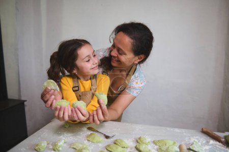 Sonriente madre amorosa y su hija feliz mirándose, sosteniendo albóndigas caseras esculpidas o varenyky ucraniano, de pie juntos en la mesa de la cocina enharinada, contra el fondo de la pared blanca
