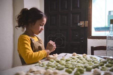 Selbstbewusstes Porträt des süßen kleinen Mädchens in beiger Schürze, das am Küchentisch mit geformten Knödeln steht, Teig füllt und Ravioli formt, ihrer Mutter beim Kochen hilft, allein das Abendessen zubereitet
