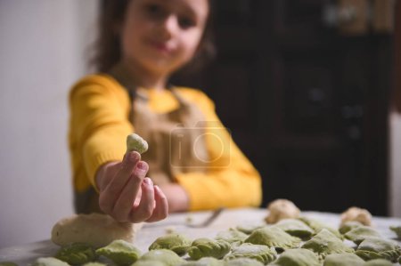 Close-up hand of a blurred child girl, little chef in beige apron, holds out at camera a homemade dumpling, smiles looking at camera, standing at floured table with molded ravioli, pelmeni, vareniki