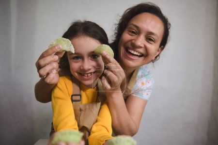 Close-up portrait of a mother and daughter having fun, smiling looking at camera, holding molded dumplings in the home kitchen. Mother and daughter cooking together