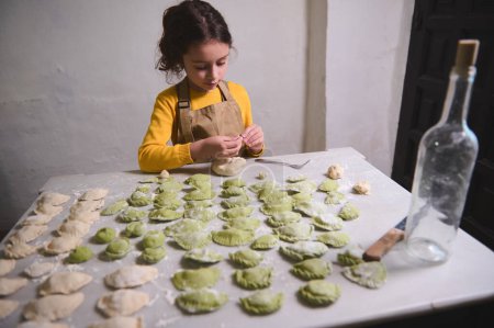 Cute little kid girl stuffing dough with mashed potatoes, modeling dumplings, standing at kitchen table with kneaded dough and ingredients. The process of cooking ravioli, varenniki, vegan pelmeni