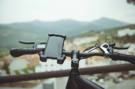 Foto de Enfoque selectivo en un smartphone fijado en el manillar de una bicicleta eléctrica moderna, con pantalla táctil digital negra mockup vacía, sobre el fondo de la ciudad medieval borrosa en las montañas. Copiar espacio - Imagen libre de derechos