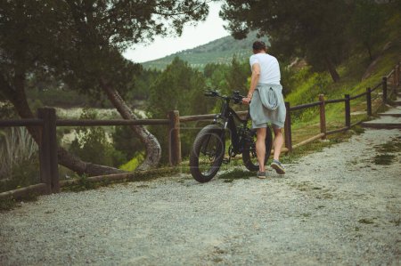 Foto de Vista trasera del joven activo arrastrando una bicicleta eléctrica en las montañas. Ciclista masculino que alquila una bicicleta a batería como modo de transporte cómodo, sostenible y ecológico - Imagen libre de derechos