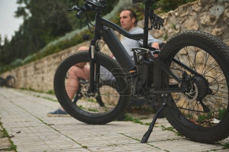 Focus sélectif sur le vélo électrique noir moderne innovant, garé près d'un banc avec un cycliste assis dans la rue de la ville. Service de partage de vélos en ville. Mobilité électronique. Personnes et mode de vie durable