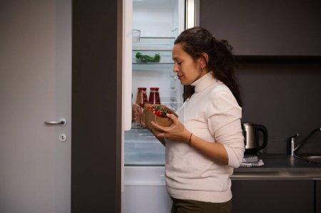 Téléchargez les photos : Femme au foyer agréable, debout au réfrigérateur à la cuisine maison, sortir une caisse en carton avec des tomates biologiques fraîches cerise, préparer une salade saine pour le dîner à l'intérieur de la cuisine maison - en image libre de droit