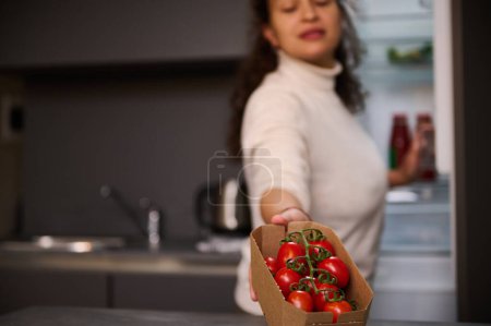 Téléchargez les photos : Une jolie femme souriante floue sort du réfrigérateur une caisse en carton recyclable avec des tomates rouges juteuses bio écologiques. Ingrédients sains pour une salade saine - en image libre de droit