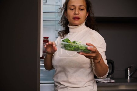 Téléchargez les photos : Une femme multi-ethnique sort une caisse en carton recyclable avec des feuilles de roquette verte bio écologique du réfrigérateur. Ingrédients sains pour une salade saine - en image libre de droit