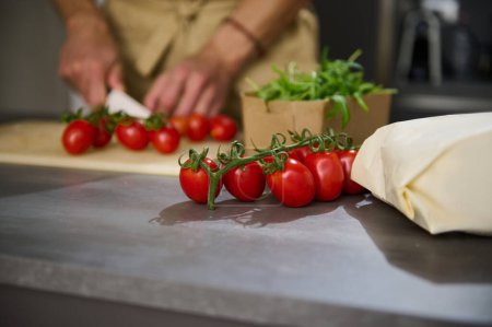Téléchargez les photos : Concentration sélective sur un bouquet de tomates rouges mûres cerise sur la table de cuisine sur le fond d'un chef masculin hachant des légumes, la cuisson de la salade pour le dîner à la maison. Espace de copie pour espace publicitaire - en image libre de droit
