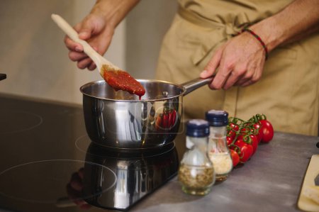 Close-up hands of a male chef holding a wooden spatula, stirring tomato juice with condiments, cooking passata according to traditional Italian recipe in the home kitchen. Mediterranean food, culinary