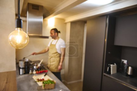 Téléchargez les photos : Focus sélectif sur une élégante lampe vintage éclairée dans l'intérieur de la cuisine moderne, contre le chef masculin flou debout à la cuisinière électrique et des ingrédients remuants, la préparation de sauce tomate traditionnelle italienne - en image libre de droit