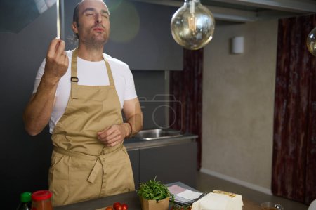 Téléchargez les photos : Jeune chef caucasien adulte dégustant des ingrédients, embrassant les doigts et faisant un délicieux geste des doigts lors de la cuisson du dîner dans la cuisine de la maison. Les gens. Cuisiner à la maison. Culinaire. Vie domestique - en image libre de droit