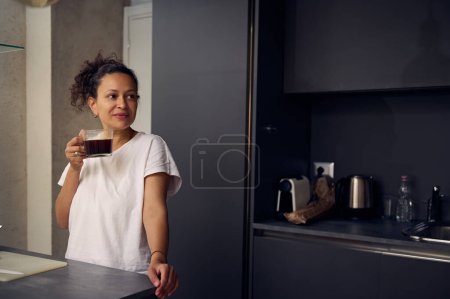 Beautiful young woman drinking freshly brewed coffee at home, smiling looking away. Multi ethnic pretty woman 40 years old, dressed in white pajamas, sipping a coffee for an energizing morning
