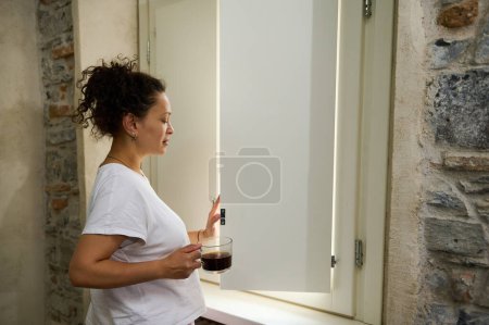 Attraktive lockige junge Frau genießt ihr Wochenende zu Hause, steht am Fenster, trinkt Kaffee und blickt verträumt aus dem Fenster