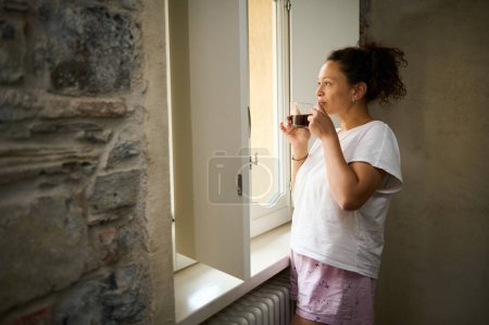 Attraktive junge erwachsene Frau im Schlafanzug, die mit einer Tasse Heißgetränk am Fenster steht, Kaffee schnuppert und die schöne Aussicht aus dem Fenster bewundert.