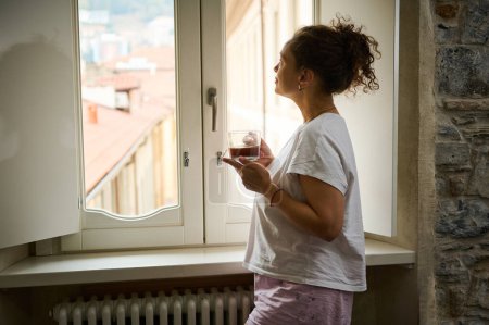 Authentisches Porträt einer schönen jungen Frau, die den schönen Ausblick auf eine mittelalterliche Stadt in Italien bewundert, während sie mit einer Tasse Heißgetränk am Fenster im Hotelzimmer steht. Menschen. Reisen. Lebensstil