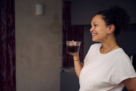 Multi ethnic curly haired young pretty woman in white pajamas t-shirt, holding a cup of freshly brewed coffee drink, smiling looking away, enjoying her happy weekend at home. Copy advertising space