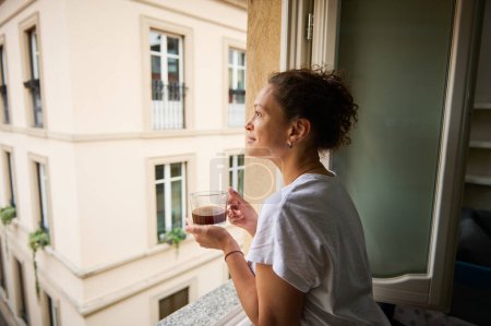 Glückliche Frau, die den Blick auf eine wunderschöne Stadt von ihrem Haus aus bewundert und am Fenster steht und am sonnigen Morgen eine Tasse Kaffee trinkt. Hübscher Tourist trinkt Kaffee und bewundert die Stadt Como